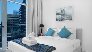 A modern hotel bedroom with a large bed, white bedding, and blue pillows. There is a framed picture on the wall and a window with a view of buildings. - Maison Privee - Beautiful Chic Apt w/Full Marina View Nxt 2 Beach (Dubai)