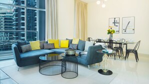 A modern living room with a sectional sofa, a dining table with chairs, and a large window offering a city view. - Maison Privee - Beautiful Chic Apt w/Full Marina View Nxt 2 Beach (Dubai)