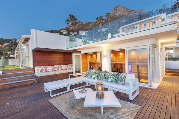 A modern outdoor living area with a wooden deck, white furniture, and a glass balustrade overlooking a mountainous landscape. - Clifton YOLO Spaces – Clifton Private Beach Villa (Cape Town)