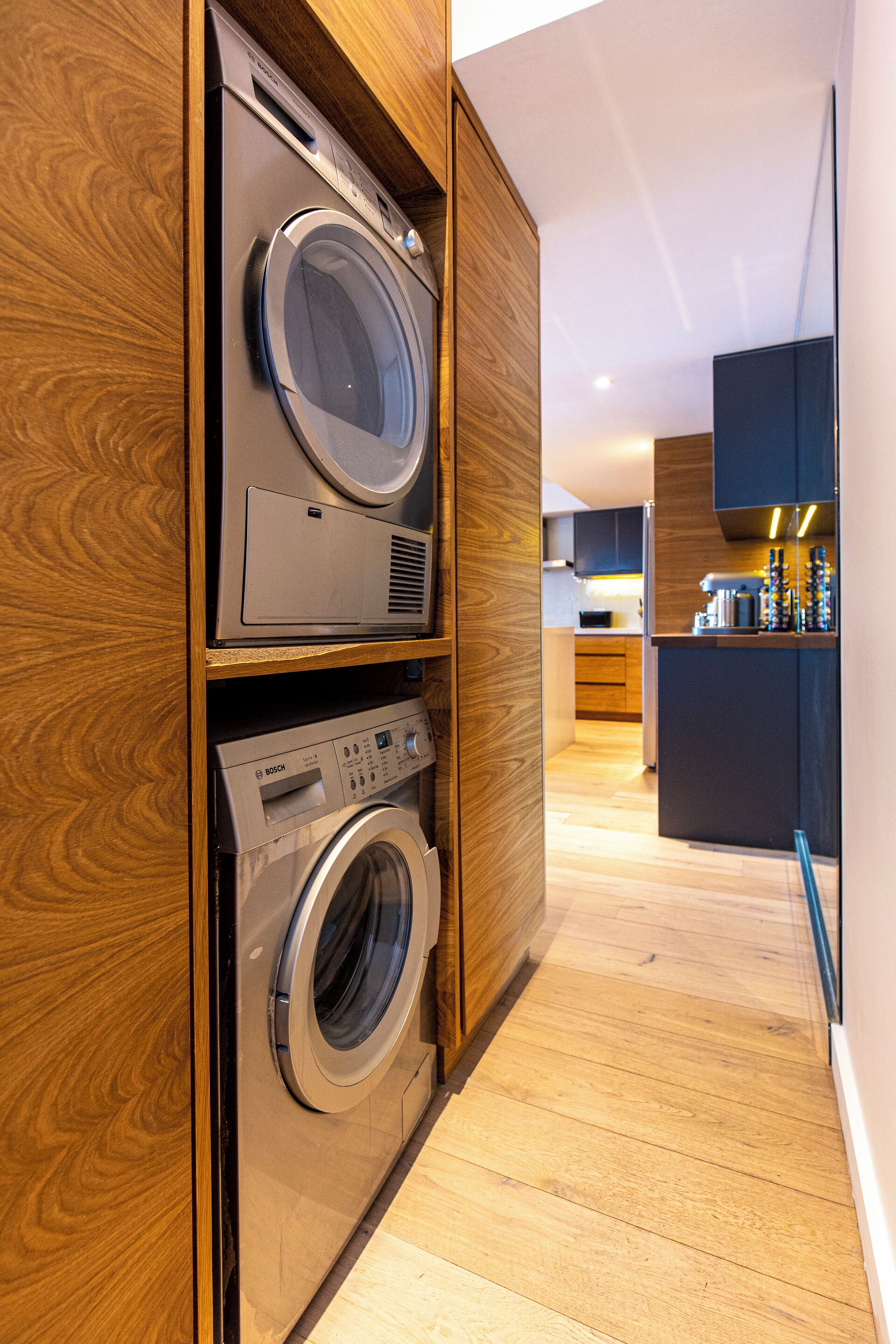 Une buanderie avec des lave-linge et des sèche-linge superposés, des meubles de rangement en bois et, au fond, un coin cuisine.