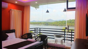 A hotel room with a bed, two chairs, a table, and a balcony with a view of a lake and mountains. - Casa Lake View Resort (Vythiri)