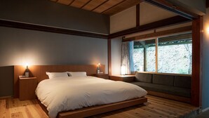 Un dormitorio con piso de madera, una cama con ropa blanca, un banco al pie de la cama y una ventana con vista a árboles.