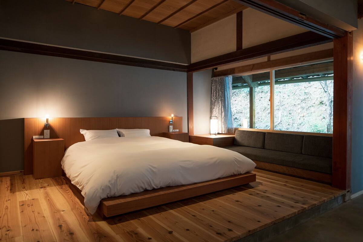 A bedroom with a wooden floor, a bed with white bedding, a bench at the foot of the bed, and a window with a view of trees.
