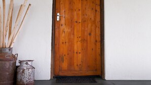 A wooden door with a metal handle, a doormat, and a potted plant with sticks. - Karoohuisie (Kopanong)