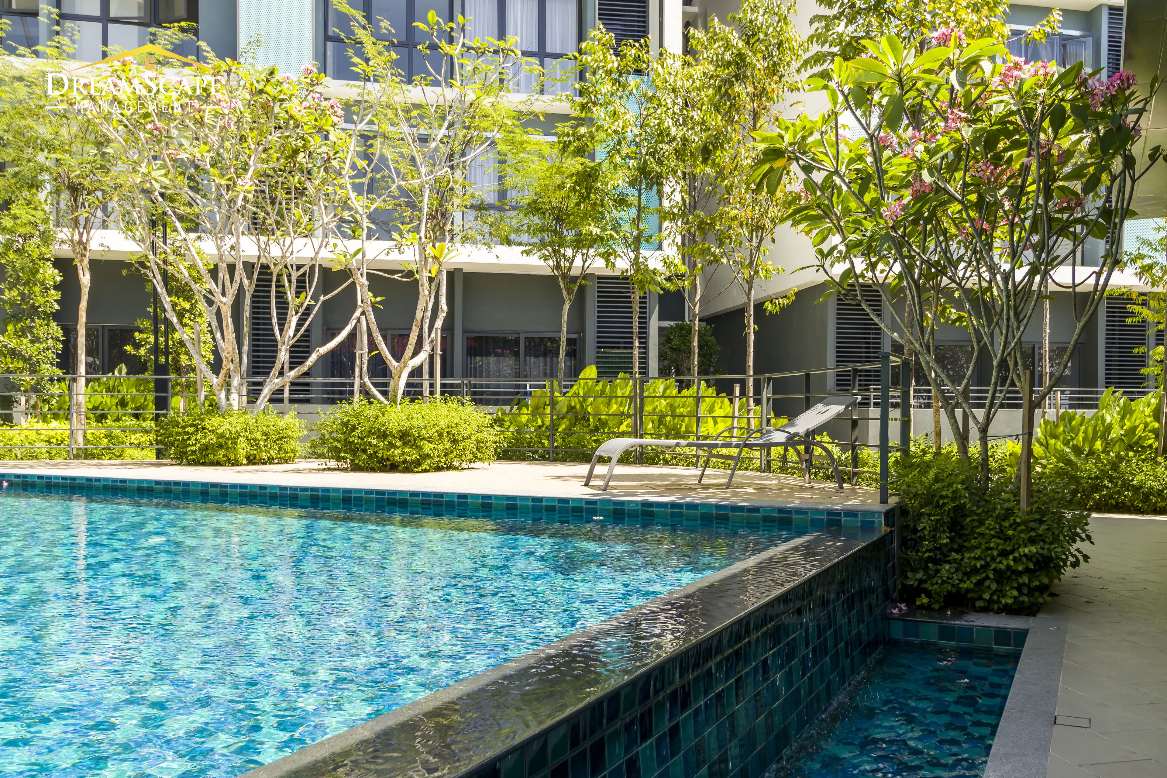 A swimming pool with a tiled edge, surrounded by greenery and a building with balconies.