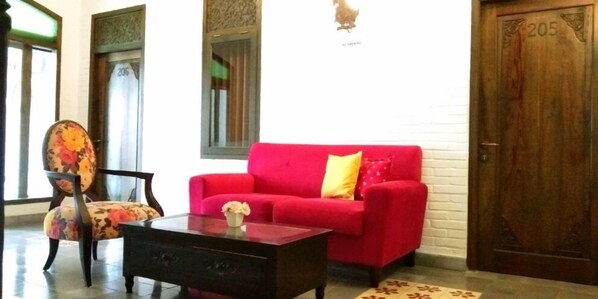 A room with a red sofa, a wooden chair, a small table with a vase, and a door marked with the number 205.
