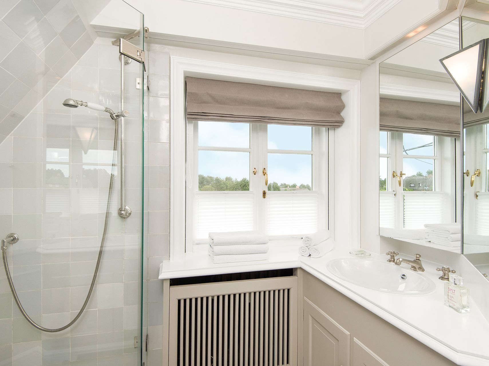 A bathroom with a glass-enclosed shower, a window with a view, and a white sink with a mirror.