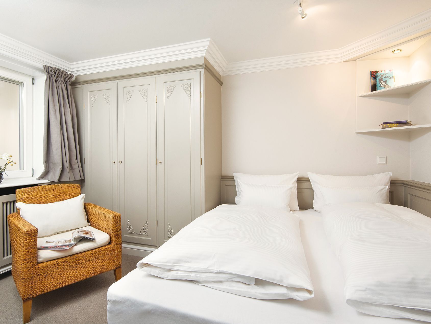 A hotel room with a bed, a wicker chair, a wardrobe, and a shelf with books.