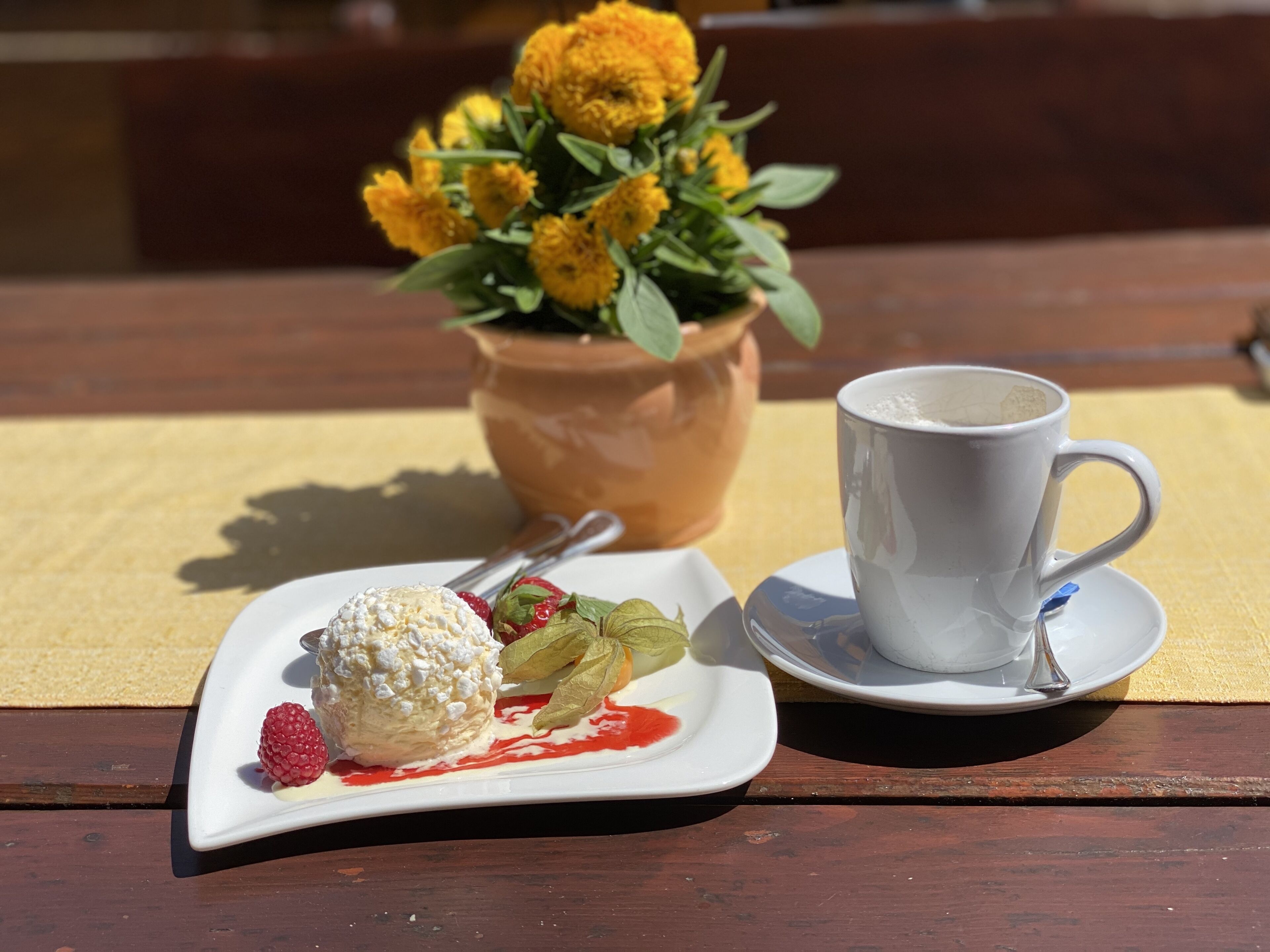 A dessert plate with a scoop of ice cream and a strawberry, a cup of coffee, and a potted plant with yellow flowers on a wooden table.