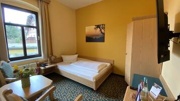 A hotel room with a bed, a desk, a chair, a window, a painting, and a wardrobe.