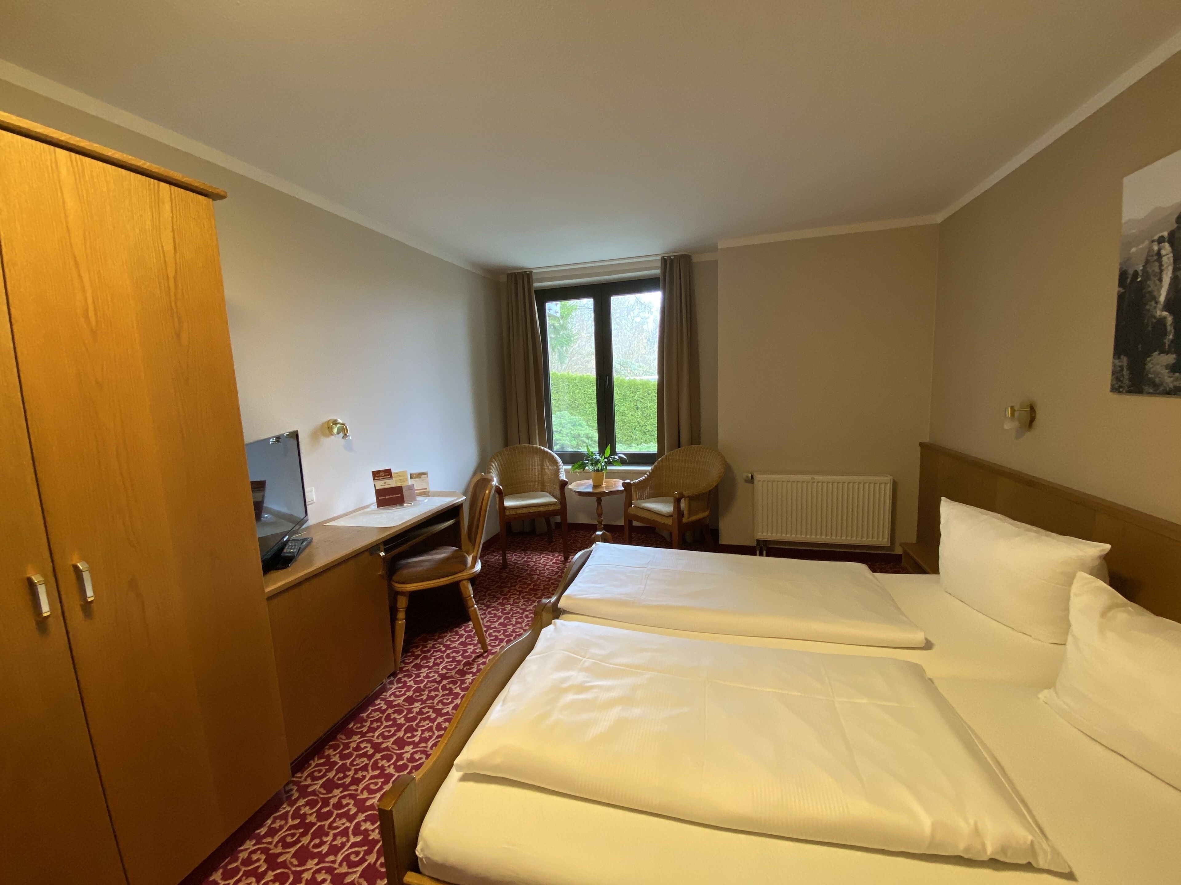 A hotel room with two beds, a desk, a chair, and a window with a view of greenery.