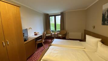 A hotel room with two beds, a desk, a chair, and a window with a view of greenery.