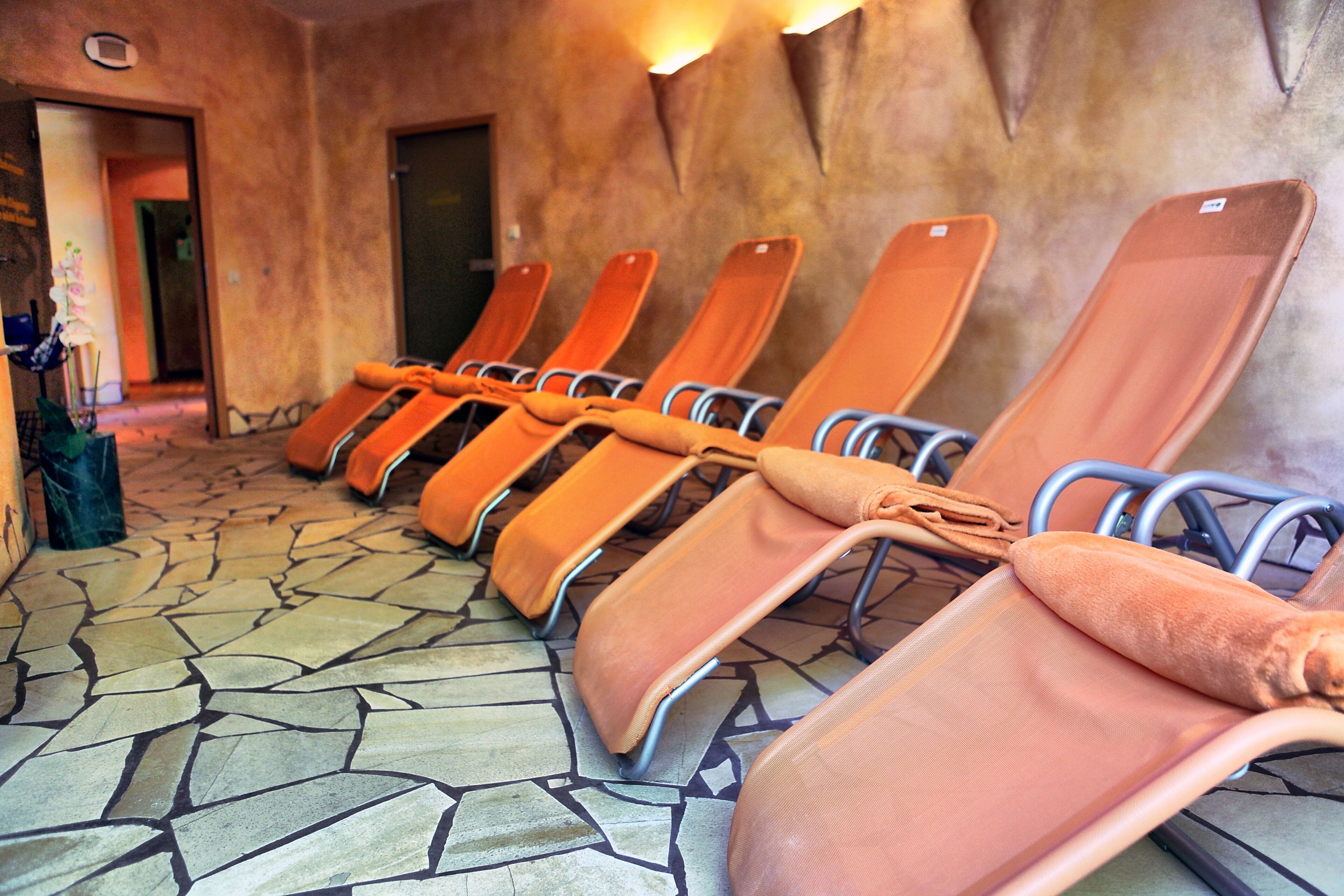 Una hilera de sillones reclinables anaranjados con estructura metálica, alineados sobre un piso de piedra estampada, en una habitación con pared texturizada y una puerta al fondo.