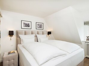 Ein ordentlich bezogenes Doppelbett mit weißen Bettwäsche, zwei an der Wand befestigte Nachttische und zwei gerahmte Bilder darüber.
