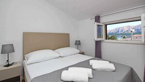A neatly made bed with white linens and a grey quilt, a bedside lamp, and a window with a mountain view. - Holiday apartment modernly furnished only 300 m from the sea (Baška)