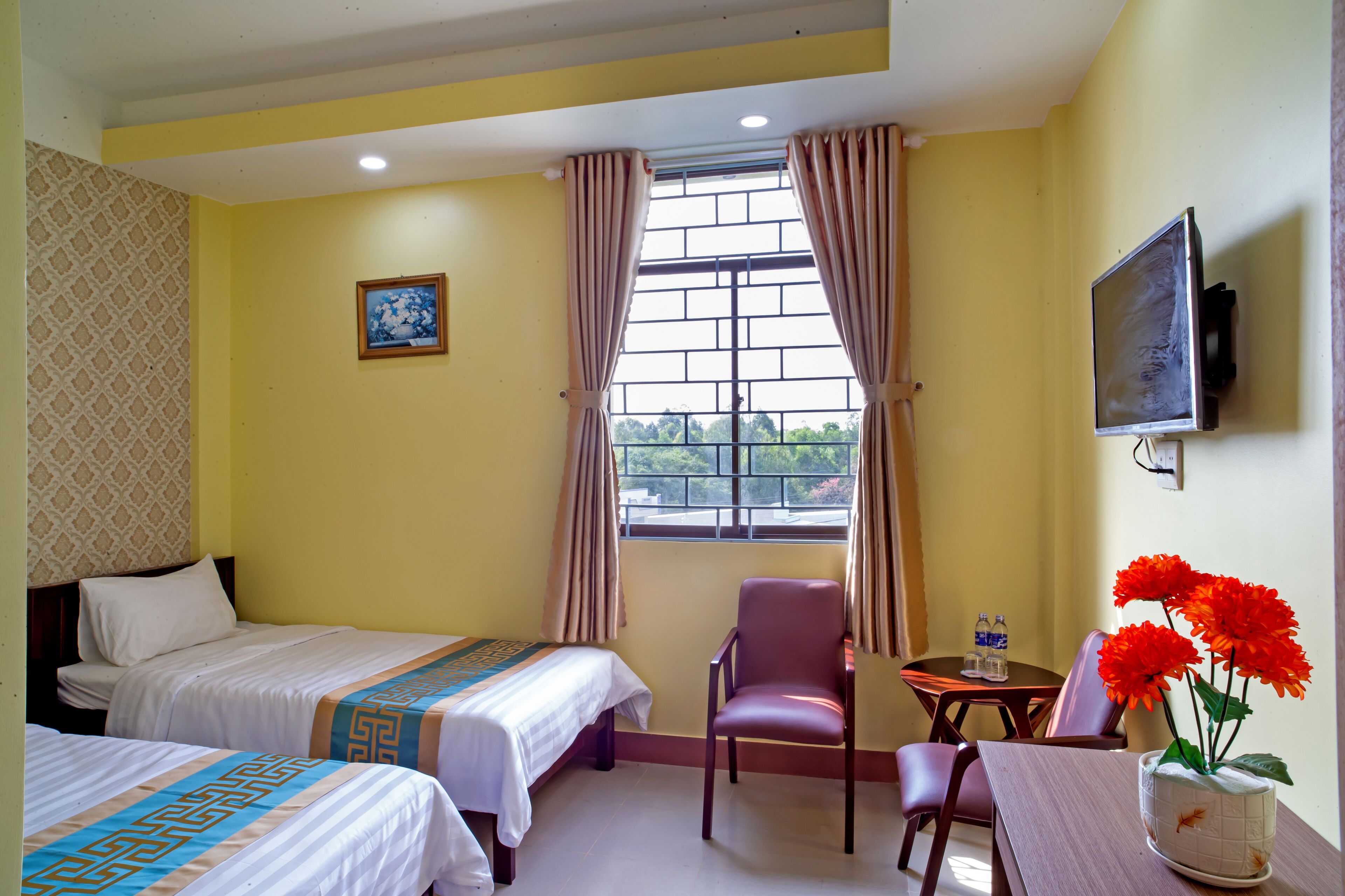 A hotel room with two beds, a chair, a small table with a vase of flowers, and a TV mounted on the wall.