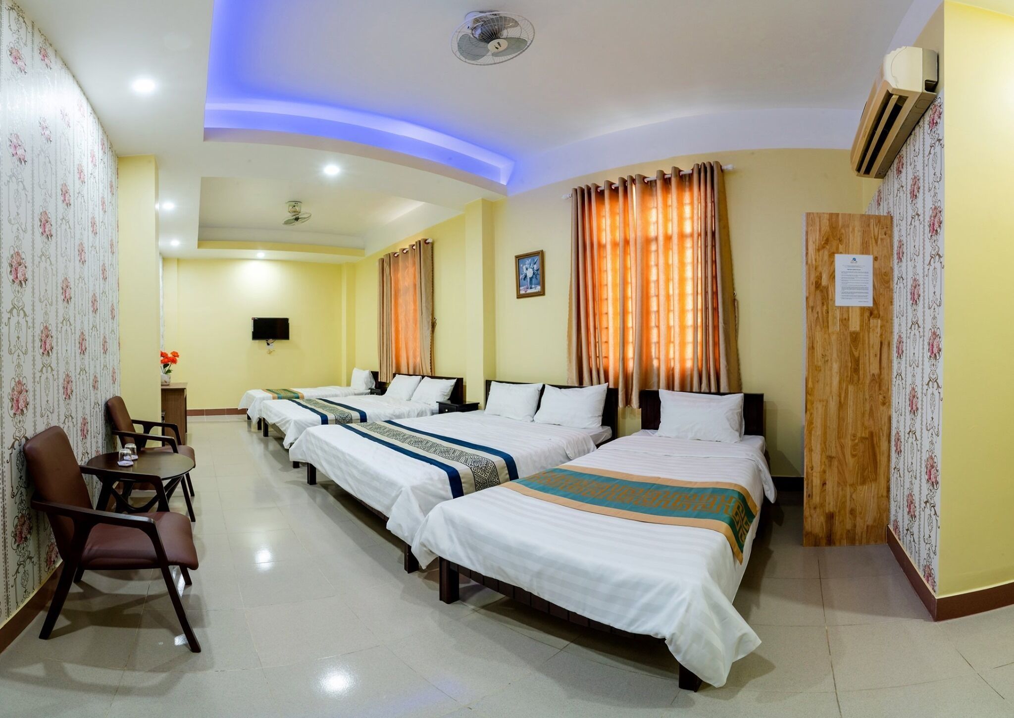 A hotel room with four single beds, a dining table with chairs, a TV, and an air conditioning unit.
