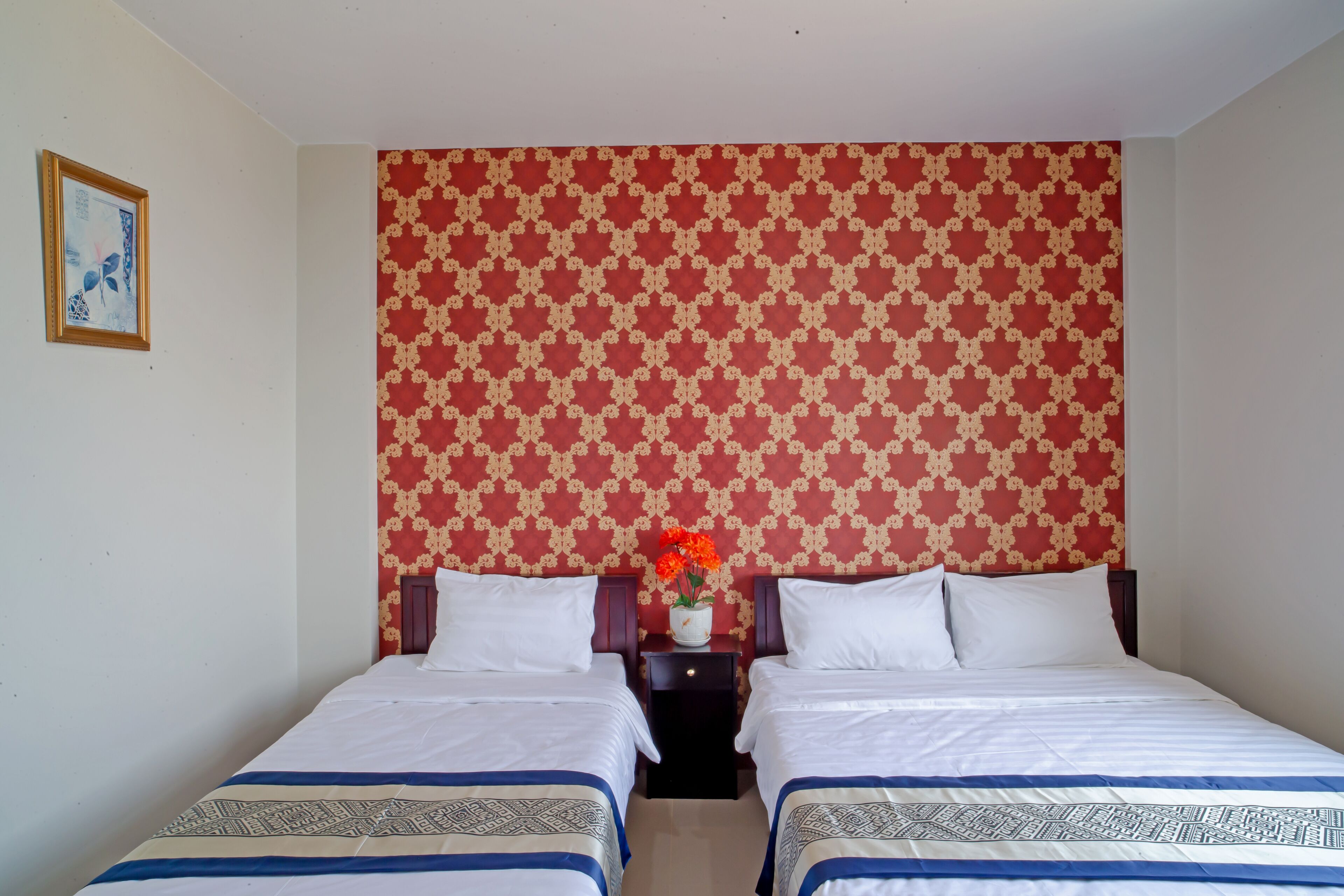 A hotel room with two beds, a patterned wallpaper, a small table with a vase of flowers, and a framed picture on the wall.