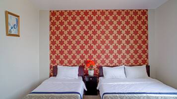 A hotel room with two beds, a patterned wallpaper, a small table with a vase of flowers, and a framed picture on the wall.
