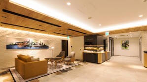 A modern lobby with a wooden ceiling, a seating area with sofas and chairs, a reception desk, and a wall with a cityscape painting.