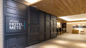 A modern hotel lobby with a dark wood panel wall, a sign reading "JR-EAST HOTEL METS", and a seating area with a yellow sofa and table.