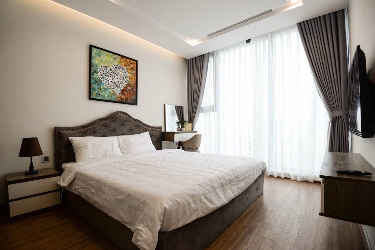A bedroom with a large bed, a nightstand, a bedside table, a desk, a chair, a window with curtains, and a wall-mounted TV.