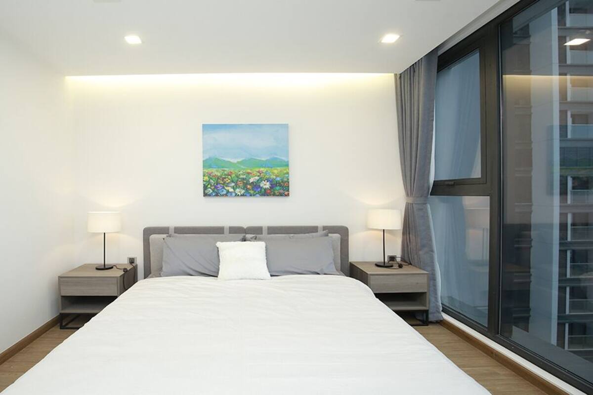A modern hotel room with a large bed, two bedside tables with lamps, a painting on the wall, and a large window with curtains.