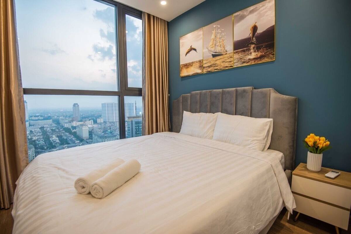 A hotel room with a large bed, a nightstand, a vase of flowers, and a cityscape view through the window.