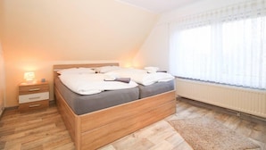 A bedroom with a wooden bed, a nightstand, a radiator, and a window with sheer curtains.