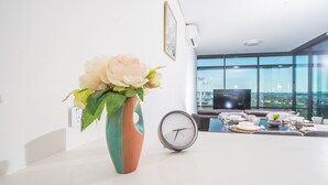 Un salon moderne avec une grande fenêtre, un vase rempli de fleurs, une horloge et un coin repas avec une table mise pour deux personnes.