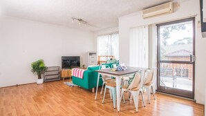 A spacious living room with a wooden floor, a dining table with chairs, a sofa, a TV, and a sliding glass door leading to an outdoor area. - Simple Comfort! 2bed1bath Unit in Meadowbank (Meadowbank)