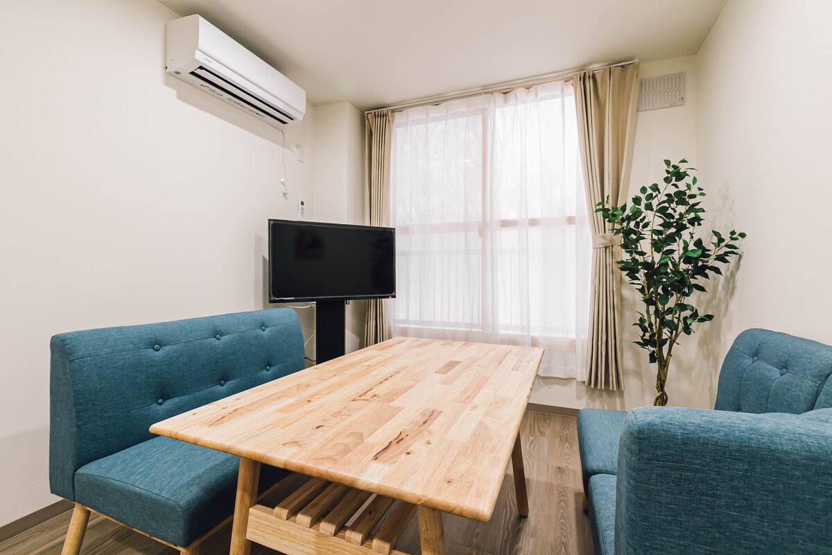 A modern living room with a wooden table, two blue sofas, a television, and a large window with curtains.