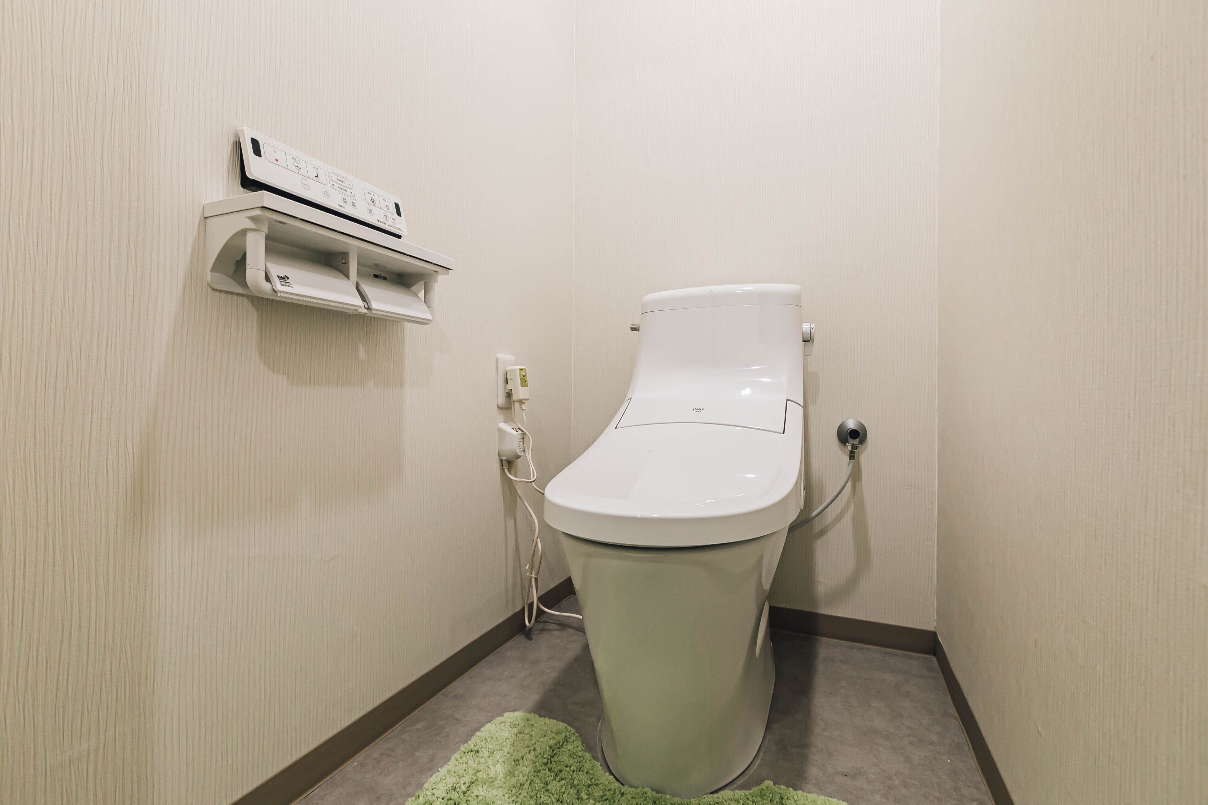 A toilet with a control panel above it, a power outlet, and a green rug on the floor.
