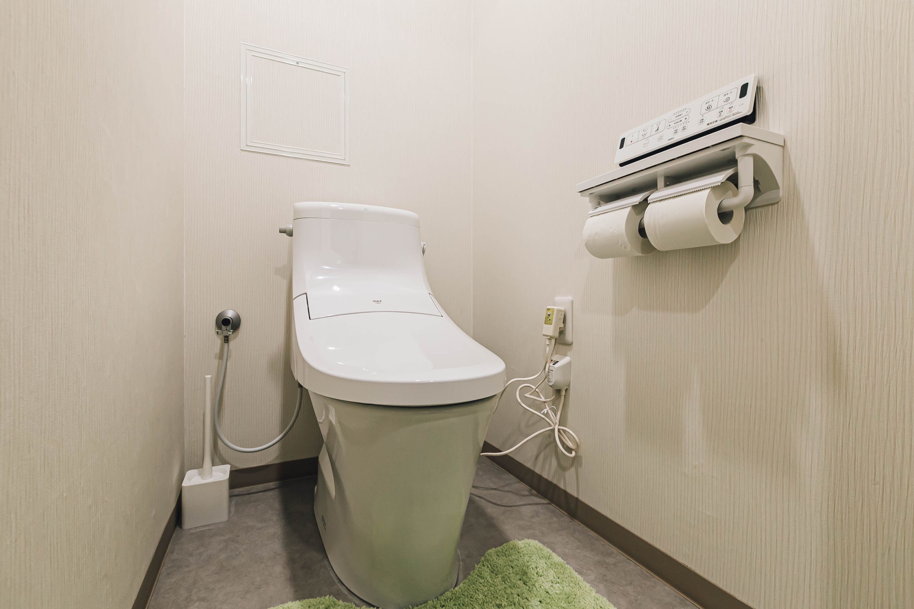 A toilet with a bidet attachment, a paper towel dispenser, and a power outlet on the wall.