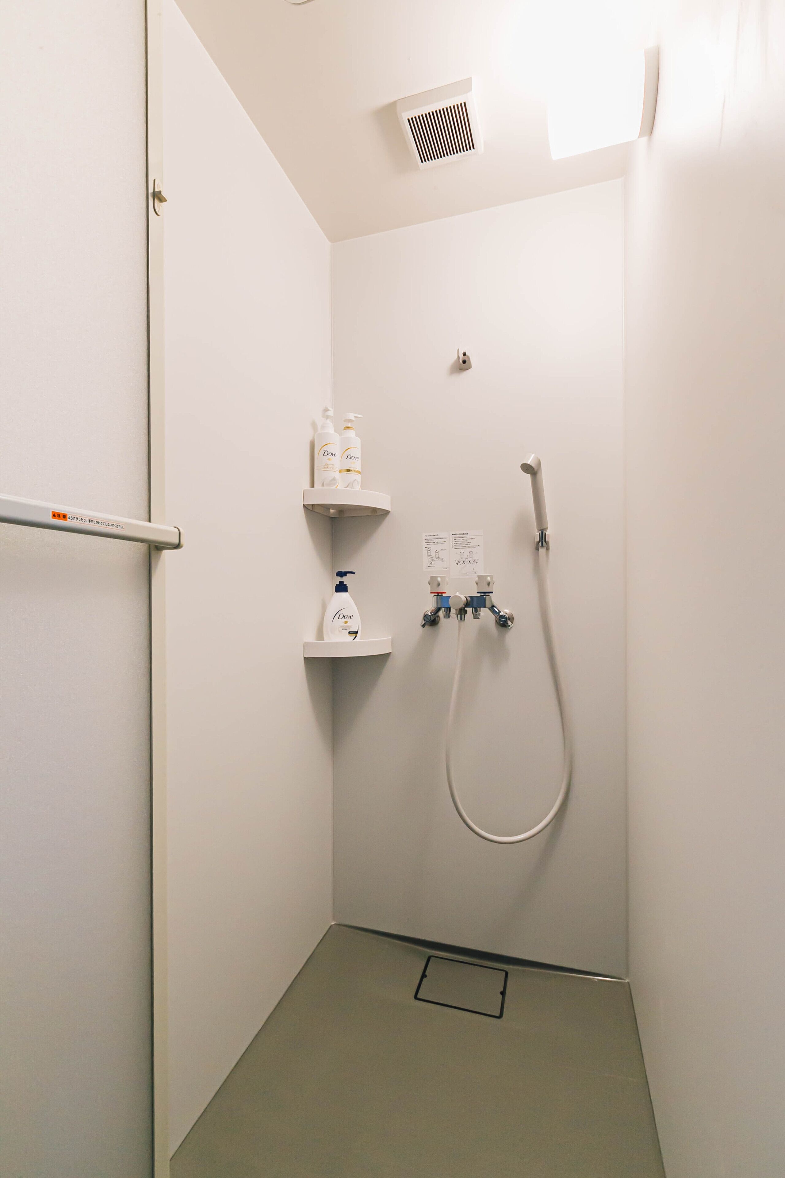 A small, white, walk-in shower with a handheld showerhead, a wall-mounted hand dryer, and a small shelf with toiletries.