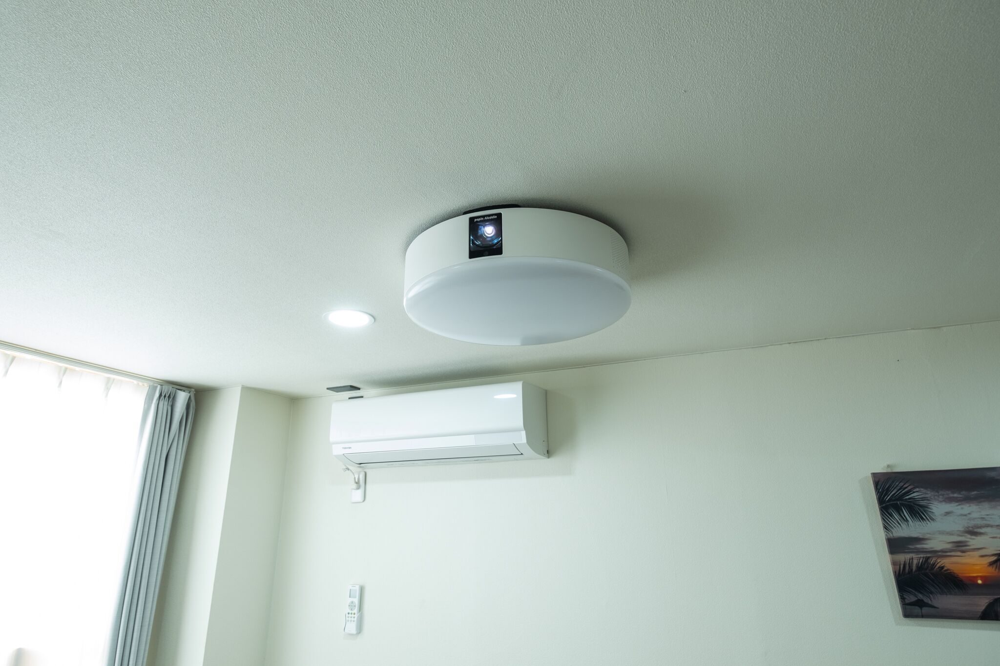 A ceiling-mounted air conditioning unit and a smoke detector in a room with a window and a wall-mounted painting.