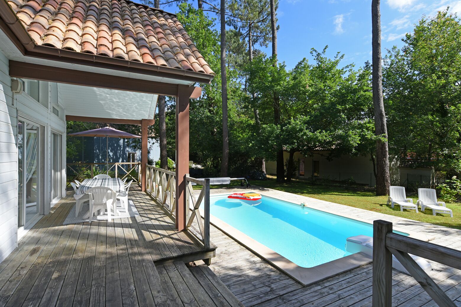 Une terrasse en bois avec une piscine, des chaises longues blanches et un espace patio couvert.