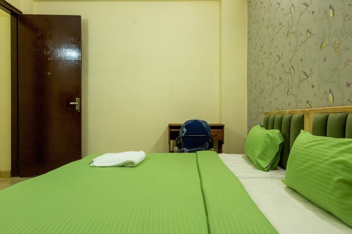 A hotel room with a green bedspread, a wooden door, a nightstand with a blue bag, and green pillows.