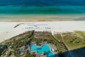 Vista aérea de un complejo turístico en la playa con una gran piscina, sombrillas y una orilla arenosa.