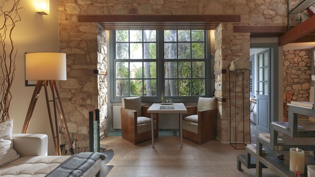 A cozy room with a stone wall, a large window, a table with chairs, and a bed with a blanket. - Live in the Acropolis' Shadow-now w/ super Terrace
 (Athina)