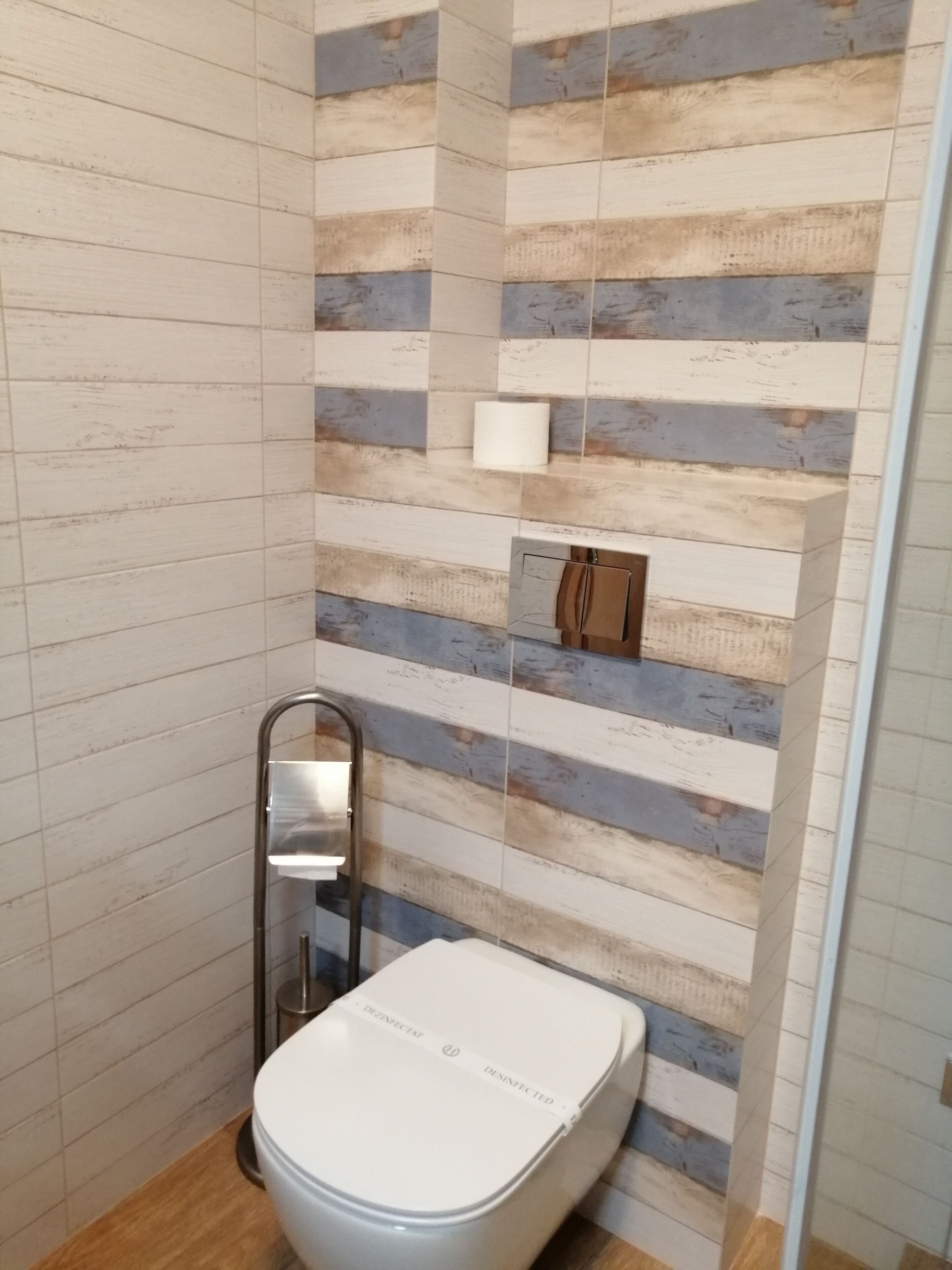 A modern bathroom with a striped tile wall, a toilet, a handrail, and a small shelf with a tissue box.