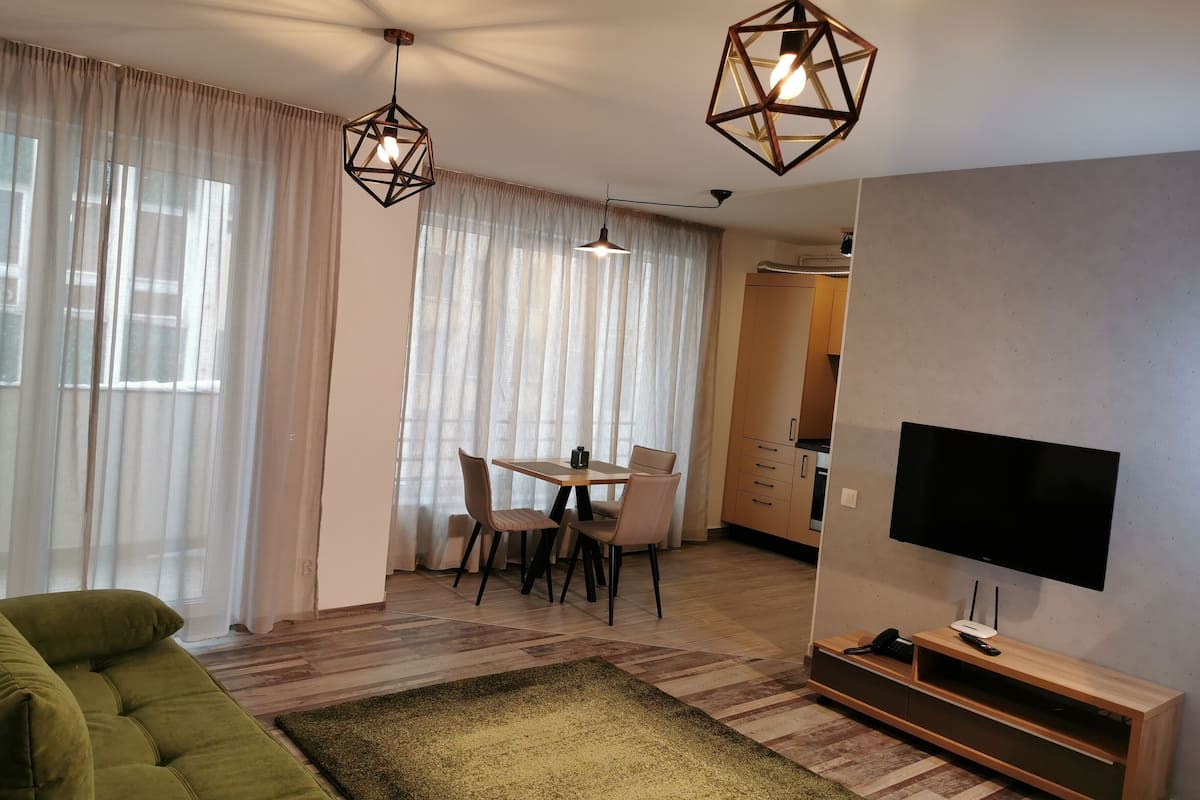 Uma sala moderna com um sofá verde, uma área de jantar com mesa e cadeiras, uma TV sobre um suporte de madeira e uma luminária pendente geométrica.