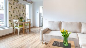 Een moderne woonkamer met een beige bank, een houten salontafel met een vaas gele bloemen, en een eethoek met een houten tafel en stoelen.