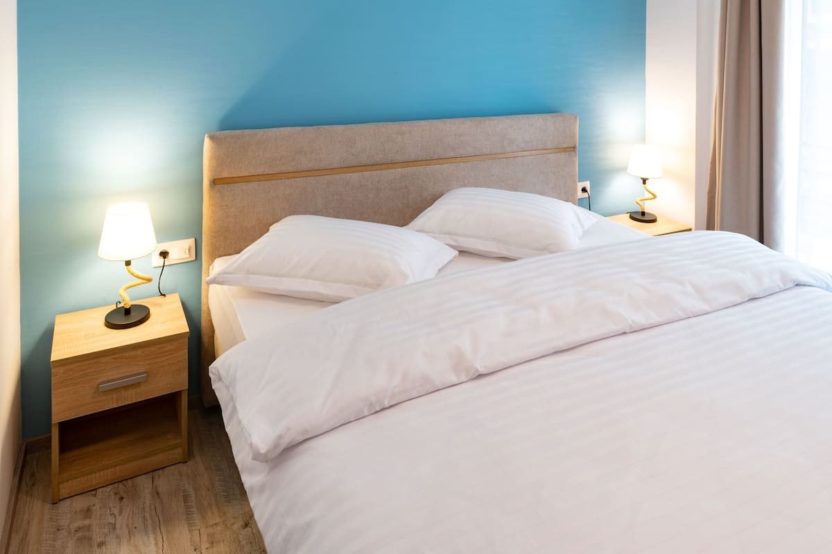 Uma cama bem arrumada com lençóis brancos, duas luminárias de cabeceira, uma mesa de cabeceira de madeira e uma parede azul.