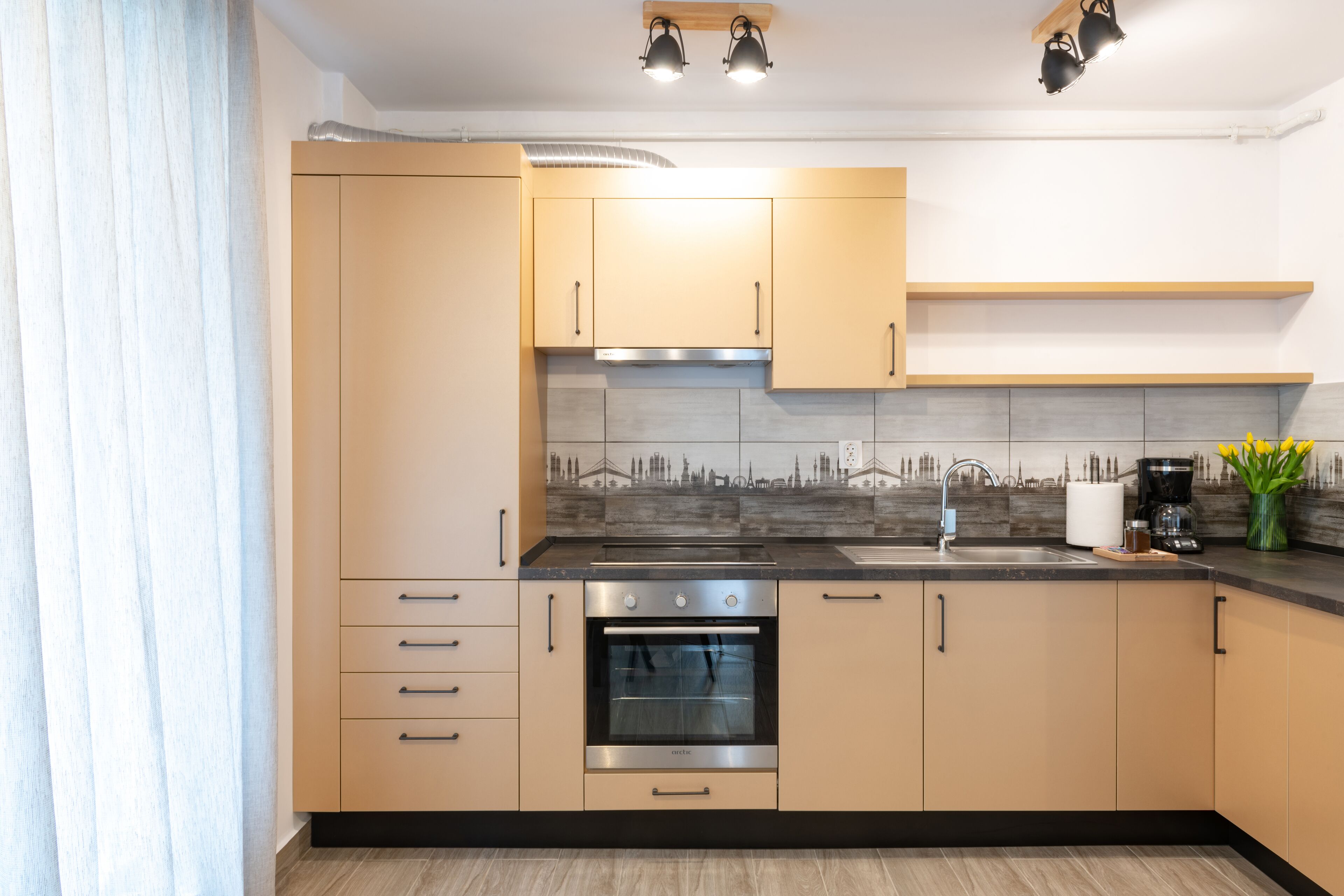 A modern kitchen with beige cabinets, a stainless steel oven, a black countertop, and a backsplash with a cityscape design.