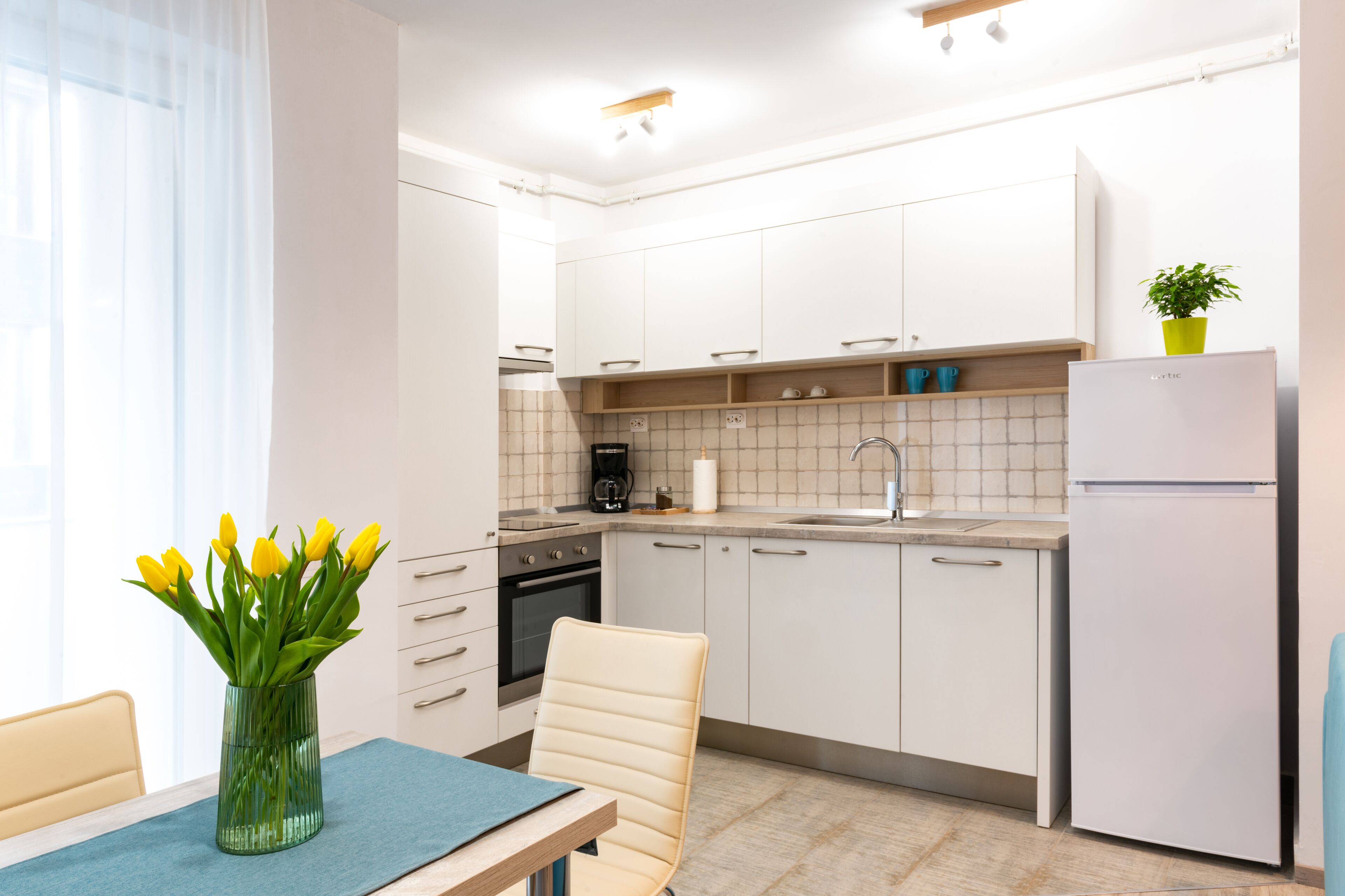 A modern kitchen with white cabinets, a built-in oven, a sink, and a refrigerator. There is a dining table with a vase of yellow flowers.