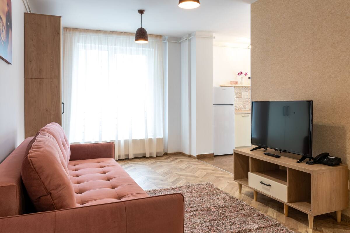 Sala de estar com sofá, televisão sobre suporte de madeira, cozinha ao fundo e janela com cortinas.