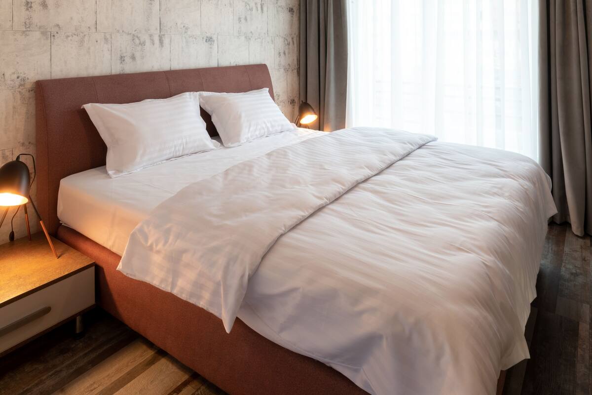 Uma cama bem arrumada com lençóis brancos, cabeceira de madeira e luminária de cabeceira.