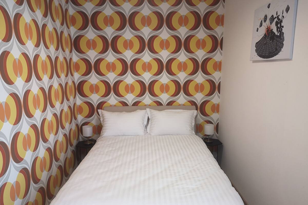 Quarto de hotel com uma cama, duas mesas de cabeceira e uma parede com papel de parede estampado em padrão circular.