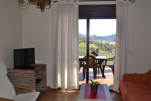 A room with a TV on a brick stand, a table with a plant, a sofa, and a balcony with a table and chairs overlooking a scenic landscape.